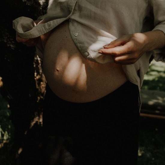 Schwangere Frau zeigt ihren Bauch, stehend vor einem Baum in der Natur.